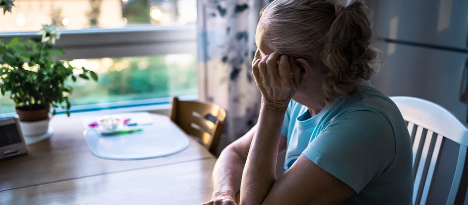 An elderly woman gazes thoughtfully out the window from her kitchen table, bathed in the soft glow of evening light, with a potted plant by her side.