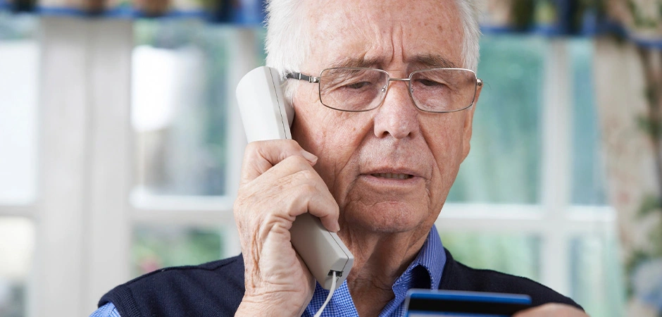 An elderly man carefully reads his credit card details while speaking on the phone, possibly conducting a financial transaction or verifying information.
