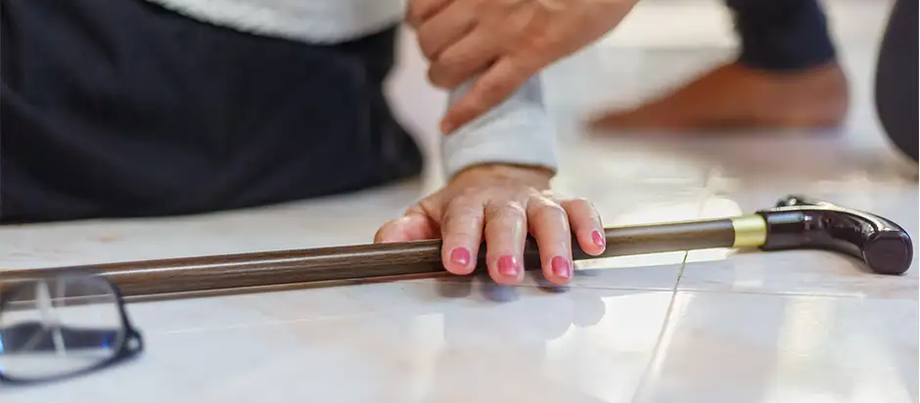 An elderly person reaches out for their cane after a fall on a tiled floor, with assistance nearby, highlighting the vulnerability and need for support in such situations.