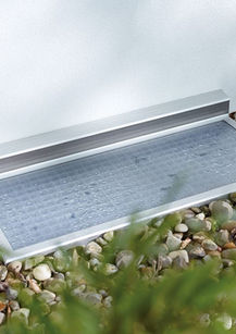 Lichtschachtabdeckung aus Aluminium mit Insektenschutzgitter auf Kiesboden.