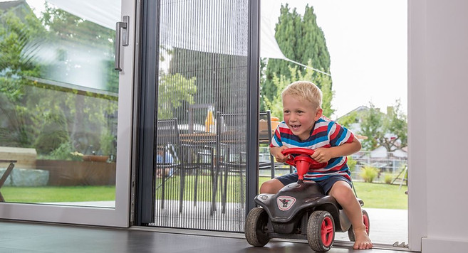 Junger Bub fährt mit Spielauto durch geöffnete Tür mit Insektenschutz-Plissee – sicherer Schutz vor Insekten und freier Zugang zugleich. Massgeschneiderte Lösungen von Bopp AG.