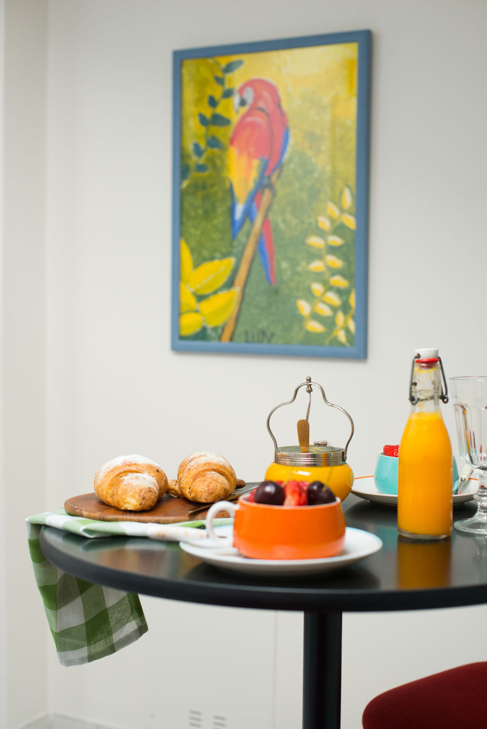 Breakfast nook with colourful parrot painting, croissants, fruit bowl, and orange juice on a black round table.