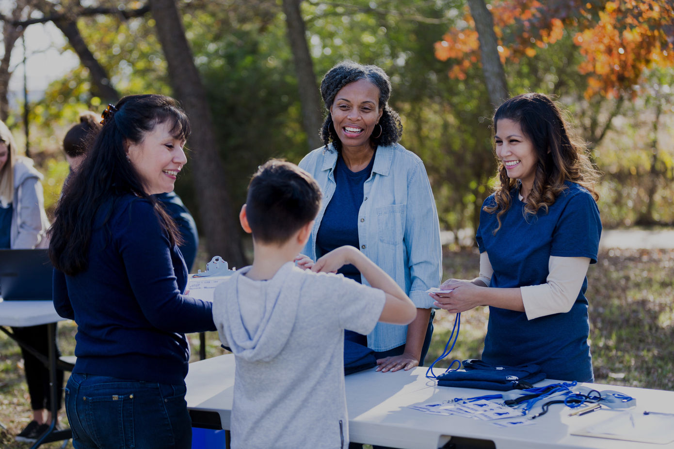 Volunteers at an outdoor information booth assisting a young person in a white shirt, with promotional materials and a blue tablecloth visible, set against an autumn backdrop.