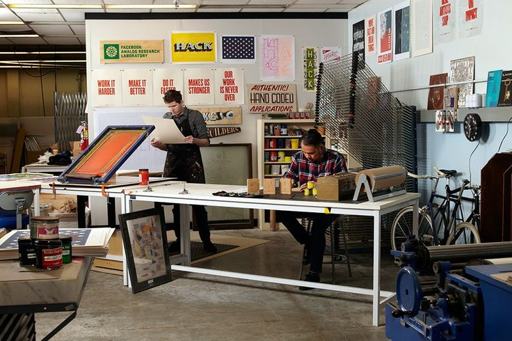 Two people work in a studio with art supplies. One examines a paper. Walls display colorful posters and text. Bicycles in the background.