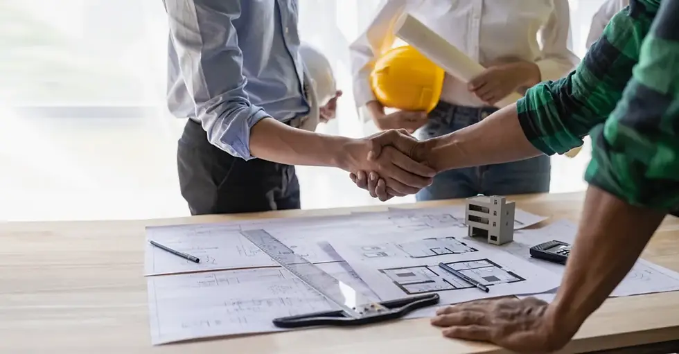Two people shake hands over house blueprints on a table while another person holds a hard hat and construction plans.