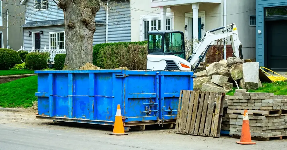A blue roll-off dumpster sits on a residential street with traffic cones around it and a stack of building materials behind it.
