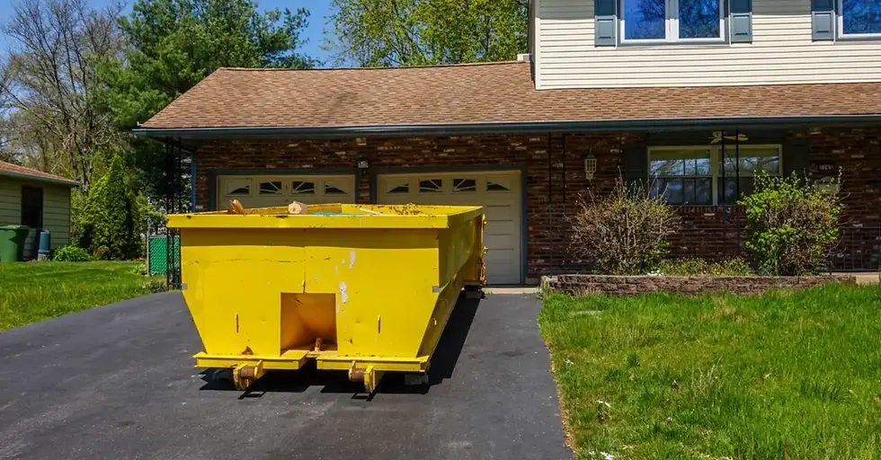 A large yellow roll-off dumpster sits in an asphalt driveway in front of a home with a lush, green lawn.