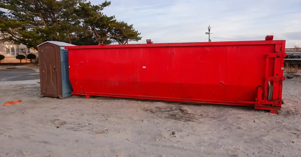 A long, red dumpster sits on a sandy beach next to a single portable restroom. The dumpster is empty.