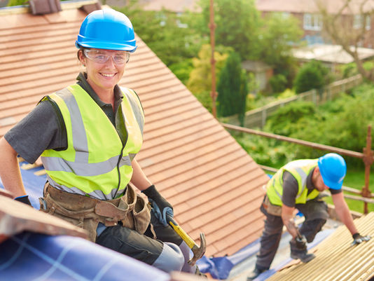 Two construction workers on a roof. One smiles, holding a hammer. Both wear blue helmets and yellow vests. Greenery in the background.