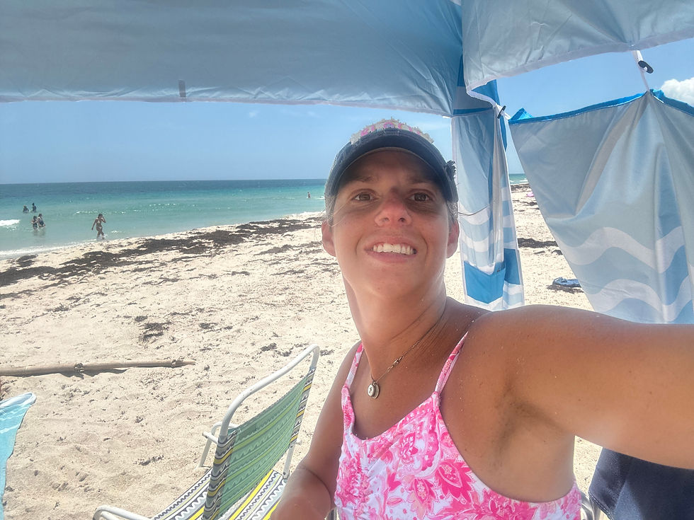 Leslie smiles for a beach selfie under a blue canopy, wearing a pink swimsuit and cap, with white sand, ocean water, and beach chairs in the background.