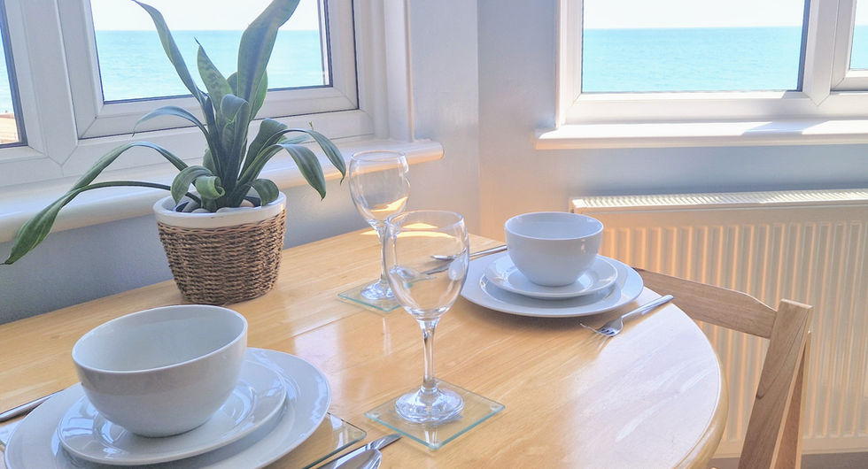 Wide view of Beachside Eastbourne Apartment 5 living area featuring a blue sofa, folding dining table, and tripe windows with sea views.