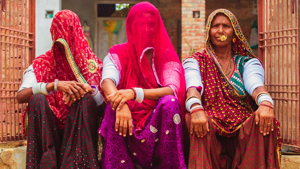 Three women in vibrant sarees sit in front of a brick building. Their faces are partially covered; the mood is serene and traditional.