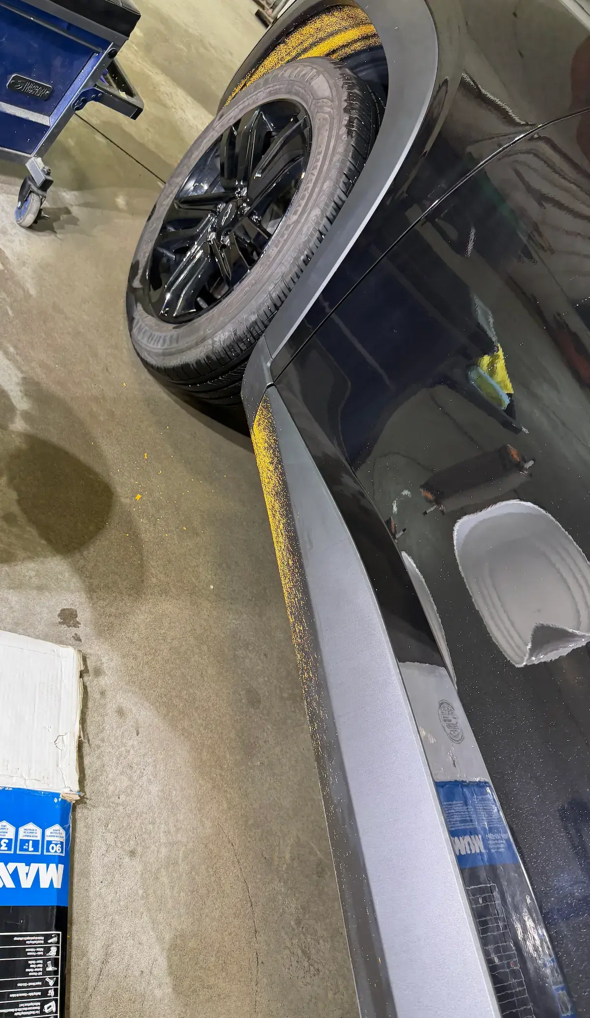 Yellow road line paint splattered across a black vehicle’s painted body panel, rocker trim and rear wheel-well area.
