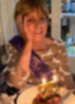 A white woman with short light colored hair sits in front of a birthday cake with lit candles in the shape of a 7 and 5