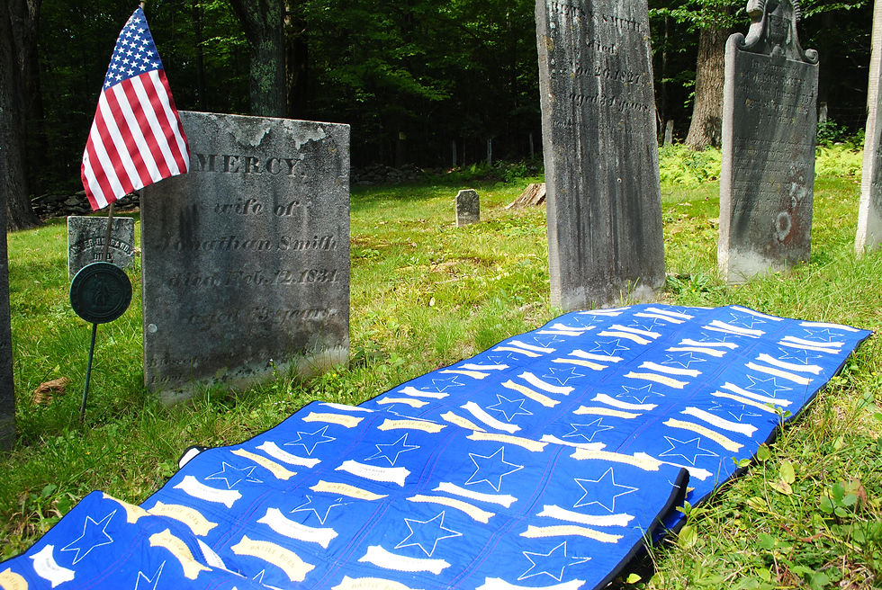 a long blue quilt splays out over bright green grass. The quilt is placed in a graveyard, as there are tombstones behind the quilt. An American flag is positioned next to the tombstone. The blue quilt is decorated with stars, and under each star is a victim's name of the Las Vegas shooting in 2017.