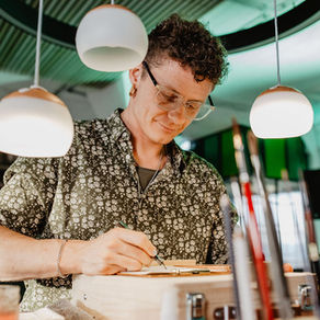 Person mit Locken und Brille (Marius) zeichnet konzentriert auf eine Holzbox in einer modernen Location unter Pendelleuchten.