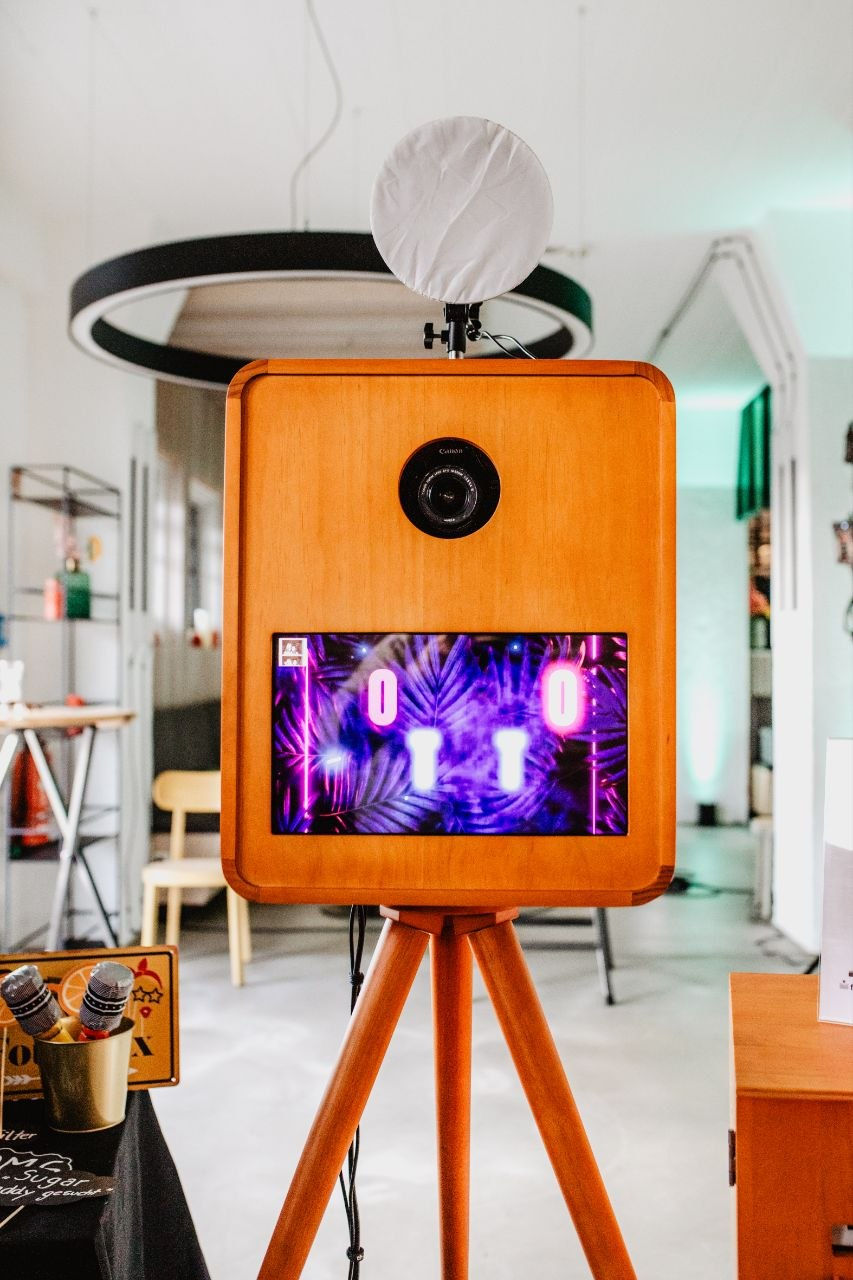 Holz-Fotobox auf Stativ in modernem Raum, mit Ringlicht und Displa in Neonfarben.