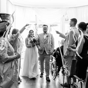 Hochzeitspaar schreitet durch ein Spalier aus Gästen in einem hellen Saal, Konfetti- und Bänderregen, freudig.