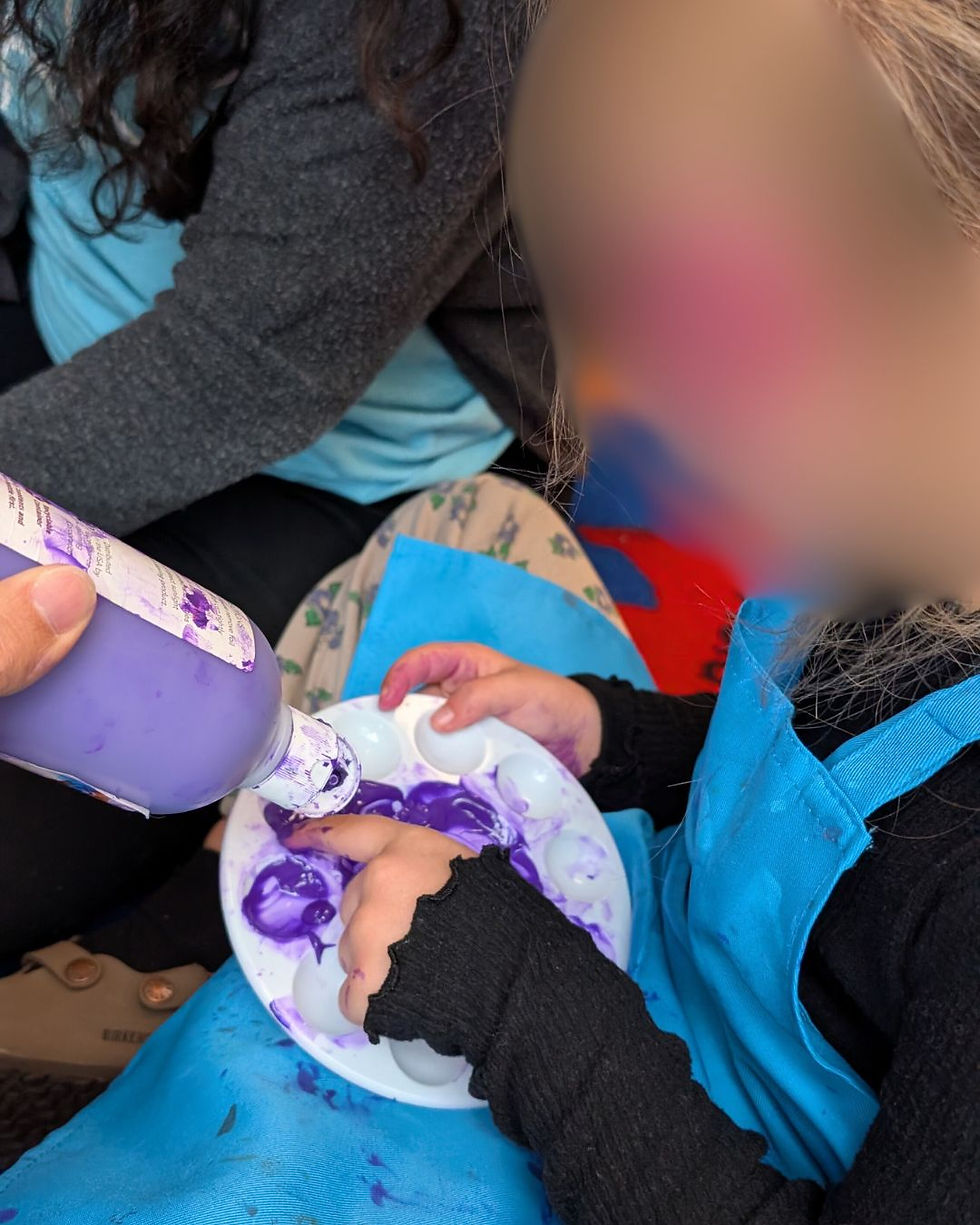 Child in a blue apron and black shirt finger-paints with purple paint on a palette. Another person helps pour more paint. Indoor art activity.