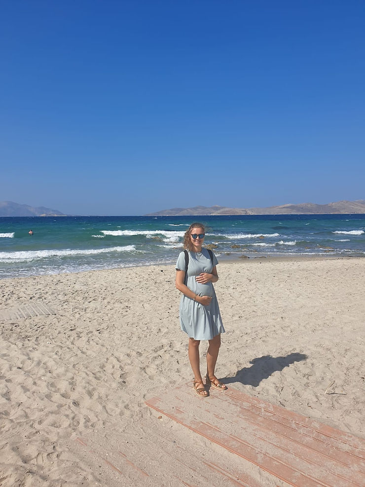Ein Sandstrand und türkises Meer. Eine Frau mit Kleid in hellblau, die ihren Babybauch hält und in die Kamera lächelt.