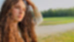 Person with curly hair gazes into distance, resting hand on head. Sunlit field and trees in background; mood is contemplative and serene.