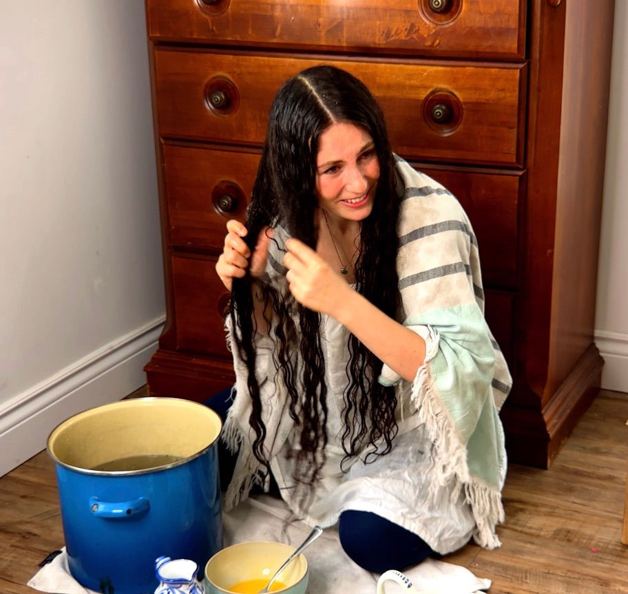 Woman with long wet curly dark hair sitting on the floor after a raw egg hair wash, holding a section of her conditioned hair next to a large blue pot during a natural Victorian-inspired hair treatment