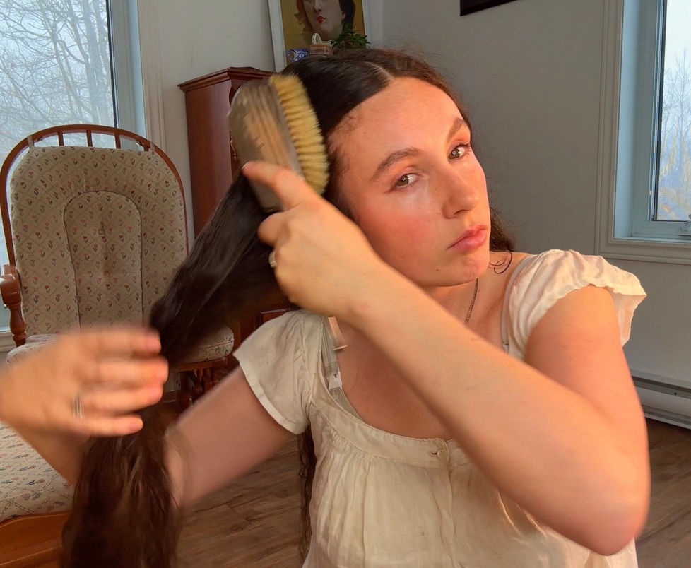 Woman with floor-length dark hair brushing thick section from roots to ends using 100% boar bristle brush in cozy room, daily Edwardian-inspired hair care routine for distributing natural oils, scalp massage, and length retention following Aline Vallandri's 1912 advice