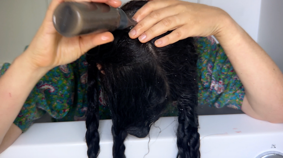 Woman applying the homemade African black soap shampoo from an applicator bottle directly onto the scalp of wet, dark braided hair while massaging it in with her fingers — demonstrating the proper application technique for the DIY natural shampoo recipe.