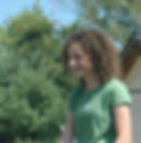 Girl with curly hair in a green shirt with a white flower, smiling outdoors. Background with trees and a building under a clear sky.