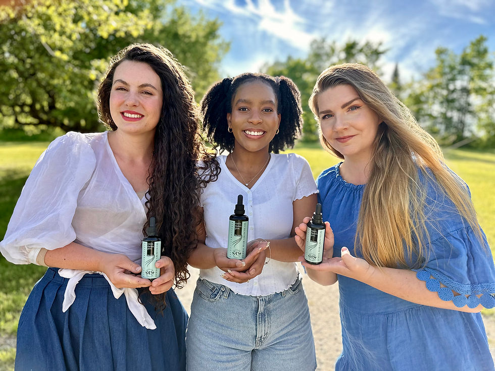 Three smiling women hold natural hair oil bottles outdoors; sunny day with trees and grass in the background. They're wearing casual, colorful outfits.