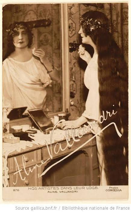 Aline Vallandri Edwardian opera singer with legendary floor-length hair, vintage 1910s postcard in her dressing room, brushing long flowing locks at vanity mirror, floral crown and elegant gown, Comœdia Paris