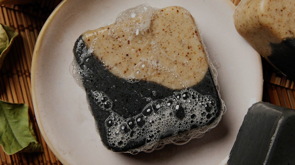 Wet square bar of layered African black soap on a ceramic plate, featuring a dark charcoal-black base and speckled beige top covered in rich white lather and bubbles, with dried leaves and bamboo mat in the background — highlighting the natural cleansing soap used as the base for the DIY shampoo recipe.