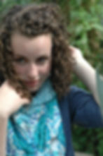 Young woman with curly hair adjusts a scarf with blue patterns, set against a leafy green background, appearing pensive.