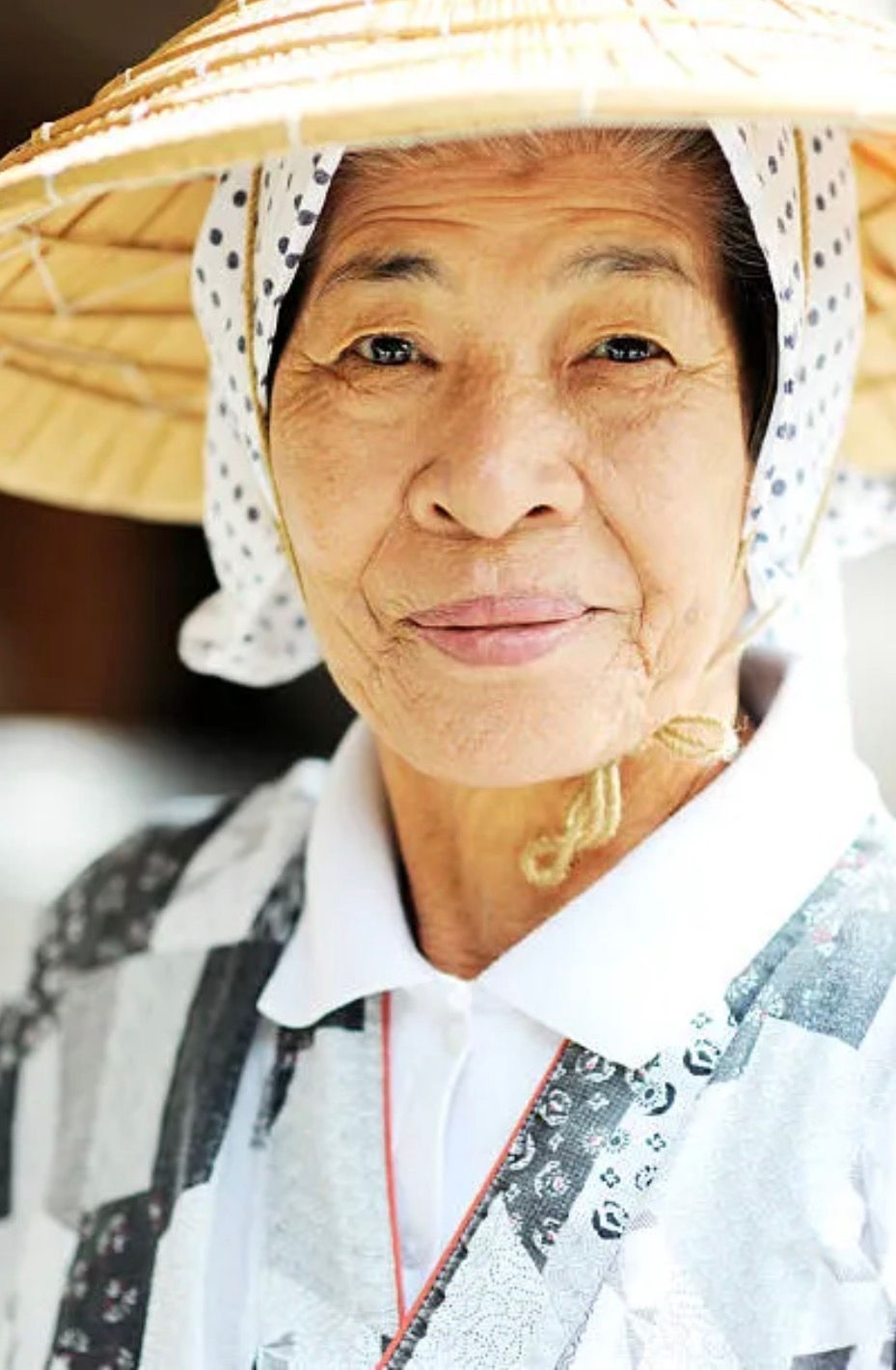 Elderly Okinawan woman wearing a traditional conical straw hat, dark hair visible underneath, smiling gently while dressed in light clothing.