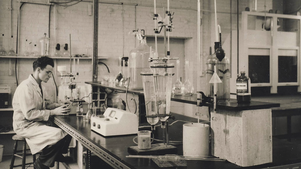 Black-and-white historical photo of a man in a lab coat seated at a laboratory bench surrounded by glassware, flasks, and scientific equipment; shelves of lab tools and a hydrogen peroxide bottle visible in the background.