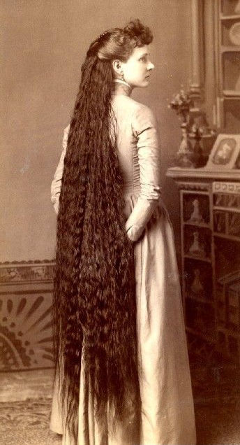 Woman with extremely long hair stands in a vintage room. Her dress is light-colored, and the setting features ornate furniture and muted tones.