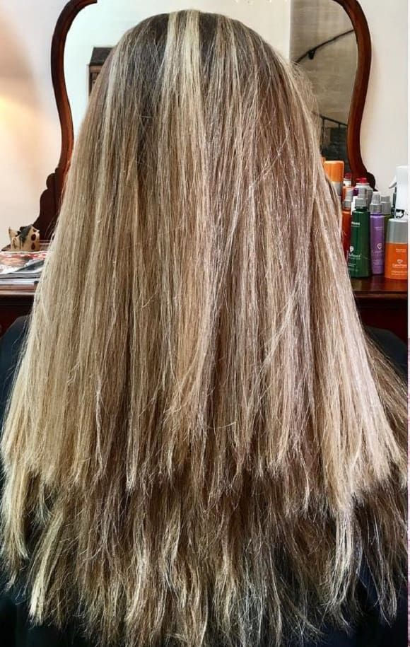 Long blonde hair with highlights in front of a mirror. Hair products visible on a shelf. Warm, inviting salon setting.