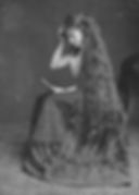 Young woman with very long hair sits on a stool, holding a book. She's wearing a patterned dress, focused, in a vintage black-and-white setting.