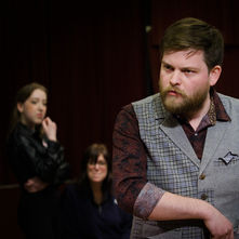A man with a beard looking to the side with other people in the background.