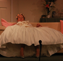 Woman in wedding dress, seated on couch, looking toward camera. Stage scene.