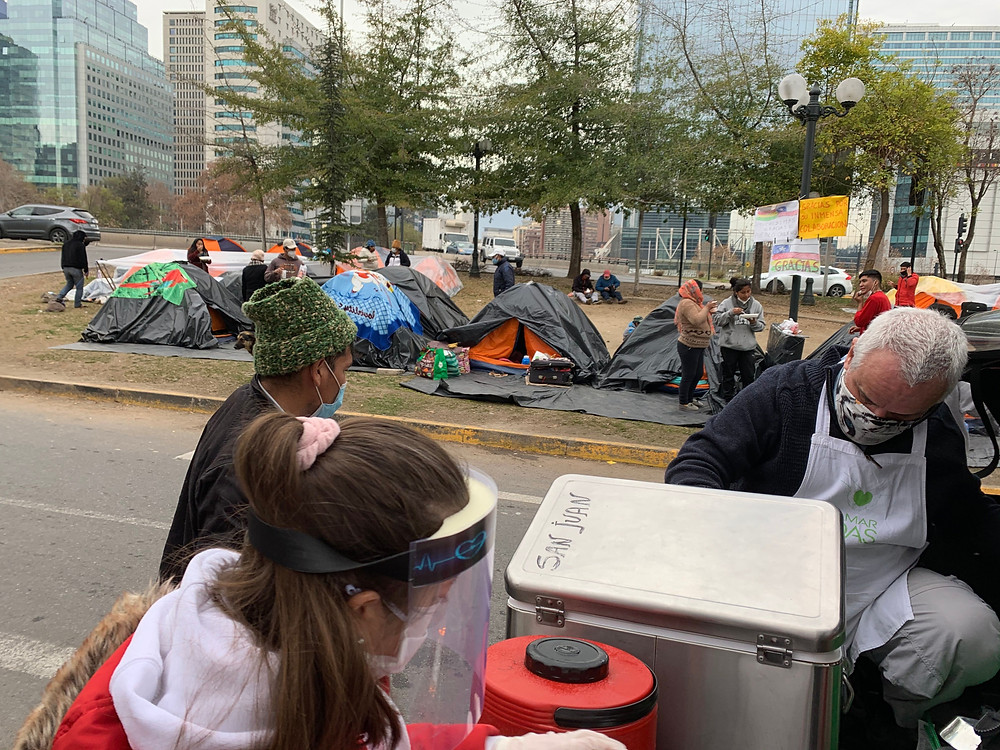people wearing masks serving food - langar - to people camping in an urban public space, in Chile.