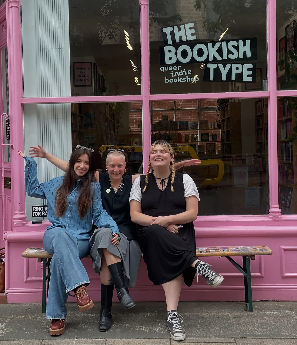 Three people smile on a bench outside a pink storefront labeled "THE BOOKISH TYPE queer indie bookshop." Casual, cheerful atmosphere.