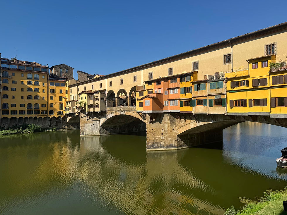 Historic bridge over river with colorful buildings. Yellow and orange facades reflect in water. Clear blue sky above, serene atmosphere.