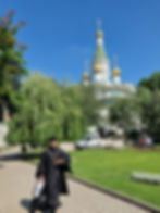 Orthodox priest in black robes walks past a church with golden domes. Sunny day, green trees, and garden workers in the background.