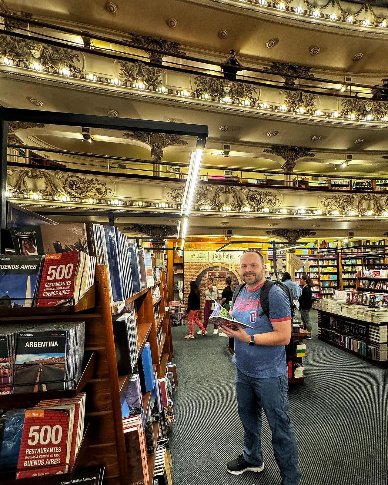 Man in blue t-shirt holds a book standing by lengthy bookshelves