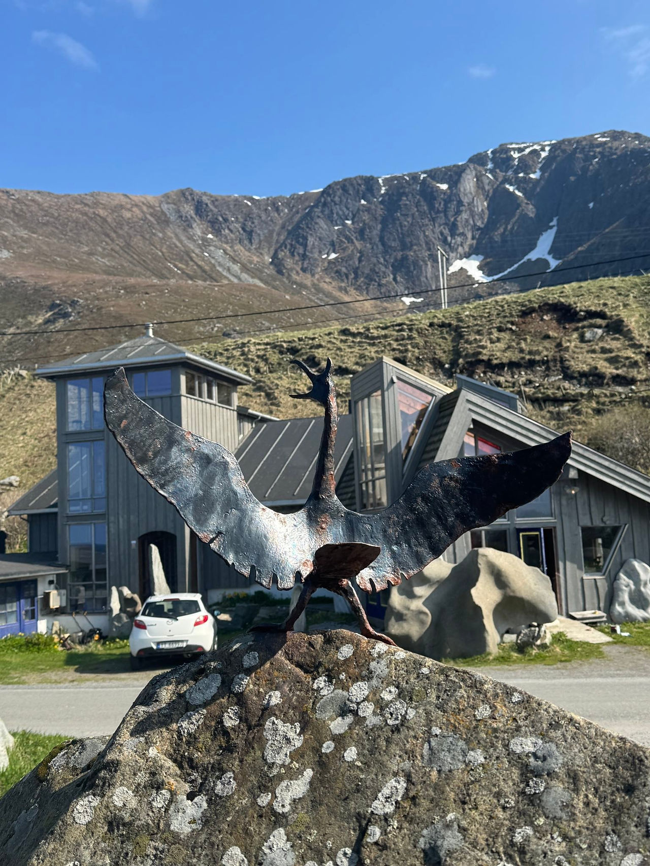 Metallic small dragon with wings outstretched against a backdrop of a house and mountain range