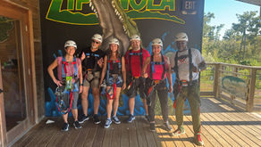 Seven people wearing white helmets and zip lining harnesses stand in front of a mural saying Zip Nola with a picture of a gator