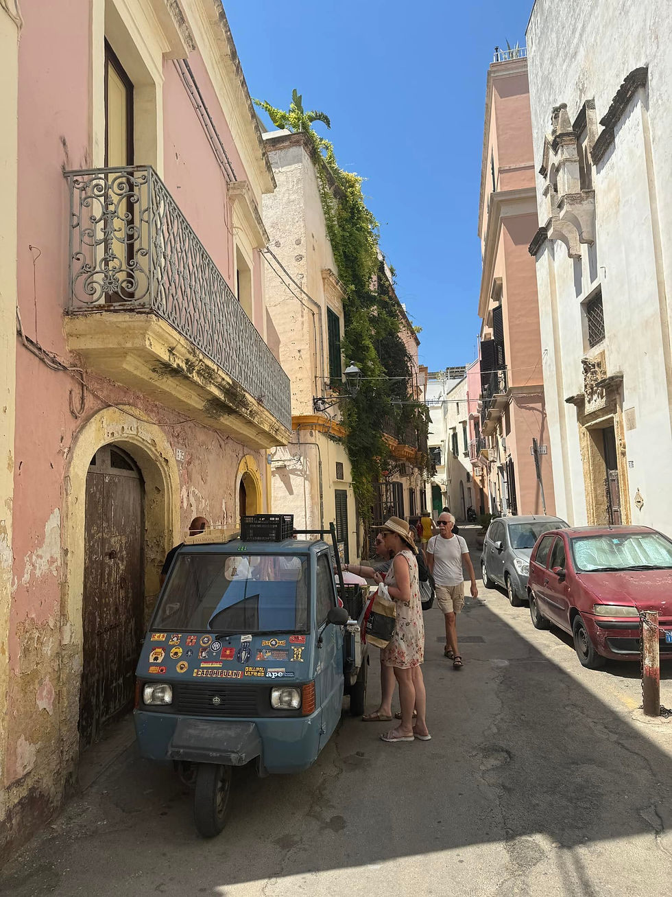 People stand by a blue tuk-tuk covered in stickers on a narrow, sunlit street lined with pastel buildings and a clear blue sky.