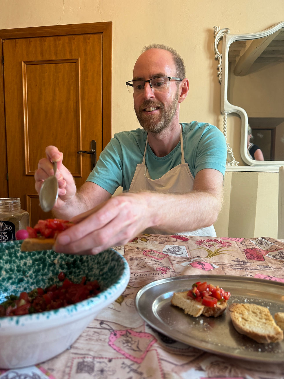 Man smiling while spreading tomato topping on bread at a kitchen table. Patterned tablecloth, wooden door, and decorative mirror in the background.