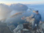 Man smiling on a mountain top with trekking pole, overlooking a coastal town and fjord at sunset. Snowy peaks in the background.